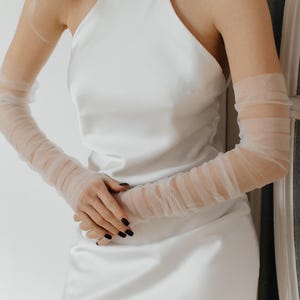 May include: A white halter top dress with a high neckline and a fitted silhouette. The dress is paired with sheer, white gloves that extend to the elbows.