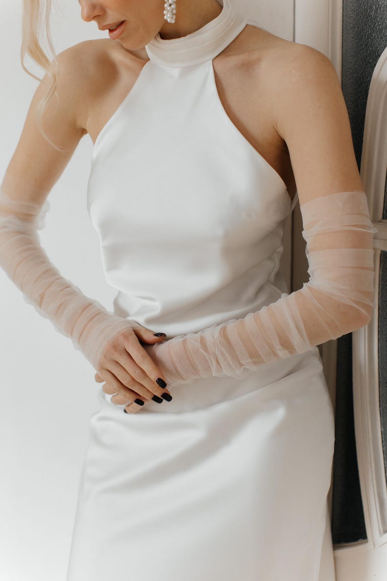 May include: A white halter top dress with a high neckline and a fitted silhouette. The dress is paired with sheer, white gloves that extend to the elbows.
