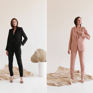 May include: Two women wearing matching suits, one in black and one in pink, standing on a beige surface. Both women are wearing heels and have their hair pulled back.