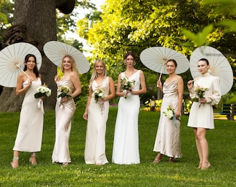 May include: Six women in white dresses stand in a garden holding white parasols and bouquets of flowers. The bride is in the center wearing a white dress with a fitted bodice and a flowing skirt. The bridesmaids are wearing white satin dresses with different styles. The women are smiling and looking at the camera.