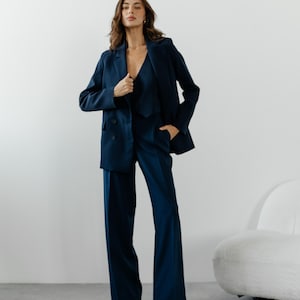 May include: A woman wearing a dark blue suit with a blazer and wide-leg pants. She is standing in front of a white wall with a white chair in the background.