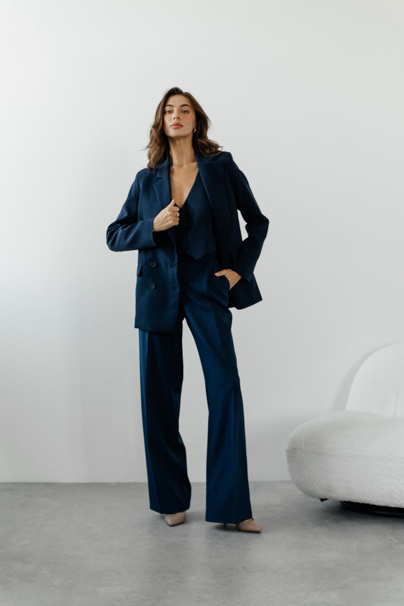 May include: A woman wearing a dark blue suit with a blazer and wide-leg pants. She is standing in front of a white wall with a white chair in the background.