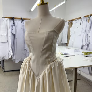 May include: A cream-colored strapless dress on a mannequin. The bodice has a structured, fitted design with a V-shaped waistline. The skirt is full and gathered. The mannequin has a gold-colored headpiece. The background shows a sewing studio.