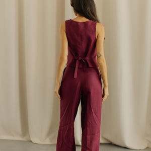 May include: A sleeveless burgundy vest with a matching pair of wide-leg pants. The vest has a tie in the back. The outfit is made of a lightweight fabric, perfect for warm weather. The model is facing away from the camera.