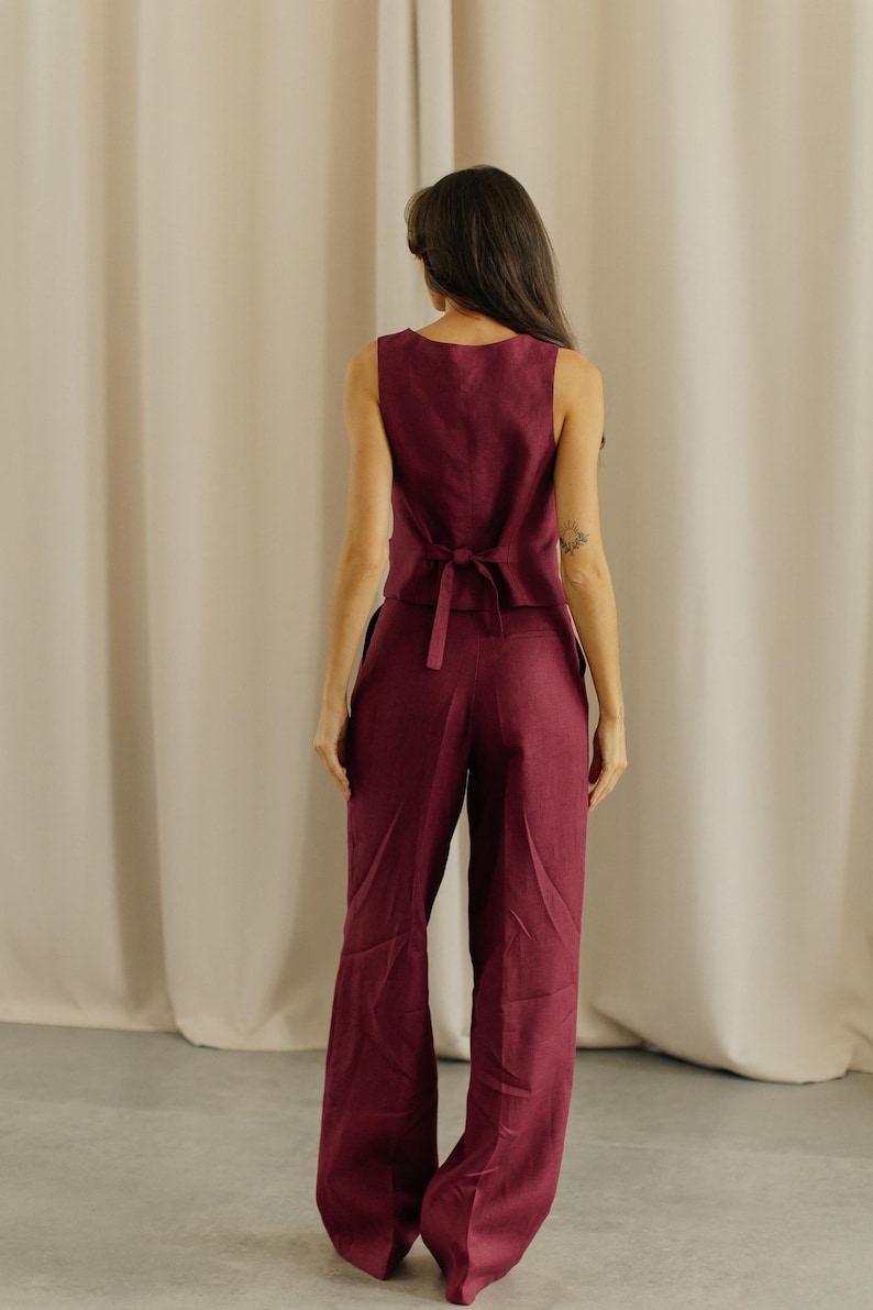 May include: A sleeveless burgundy vest with a matching pair of wide-leg pants. The vest has a tie in the back. The outfit is made of a lightweight fabric, perfect for warm weather. The model is facing away from the camera.