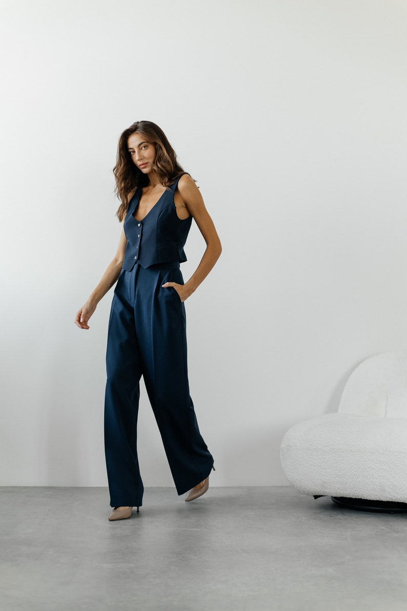 May include: A woman wearing a navy blue sleeveless vest and matching wide-leg pants. She is standing in front of a white wall and a white fluffy chair.