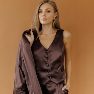 May include: A brown satin suit, consisting of a vest with buttons, matching pants, and a blazer draped over the shoulder. The outfit is a solid color and appears to be made of a smooth, shiny fabric. The background is a solid, warm brown.