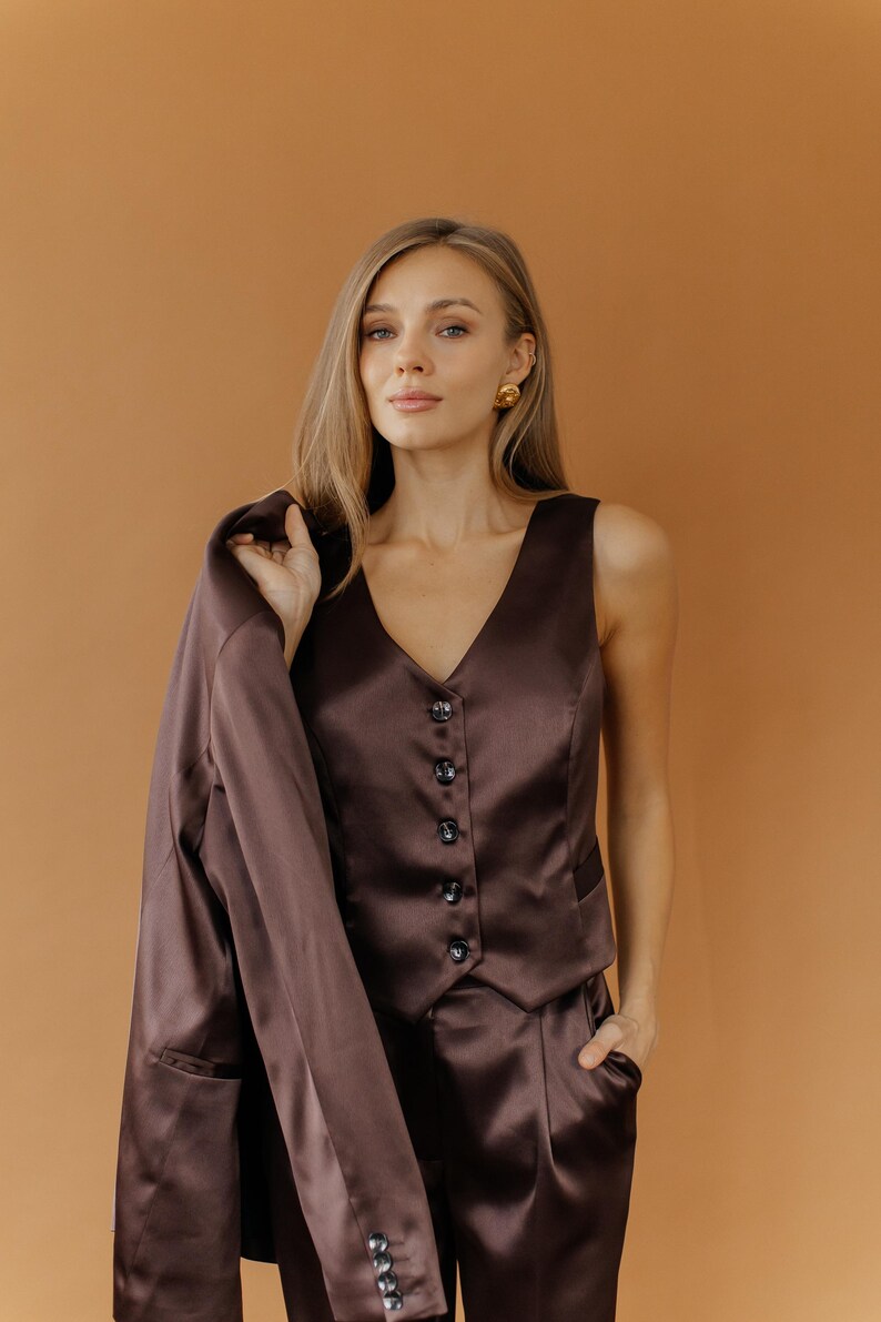May include: A brown satin suit, consisting of a vest with buttons, matching pants, and a blazer draped over the shoulder. The outfit is a solid color and appears to be made of a smooth, shiny fabric. The background is a solid, warm brown.