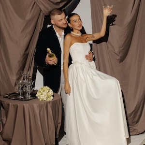 May include: A couple celebrating, with the woman in a white strapless gown raising a champagne glass. The man holds a champagne bottle. A table with champagne glasses and flowers is in the background. Brown curtains frame the scene.