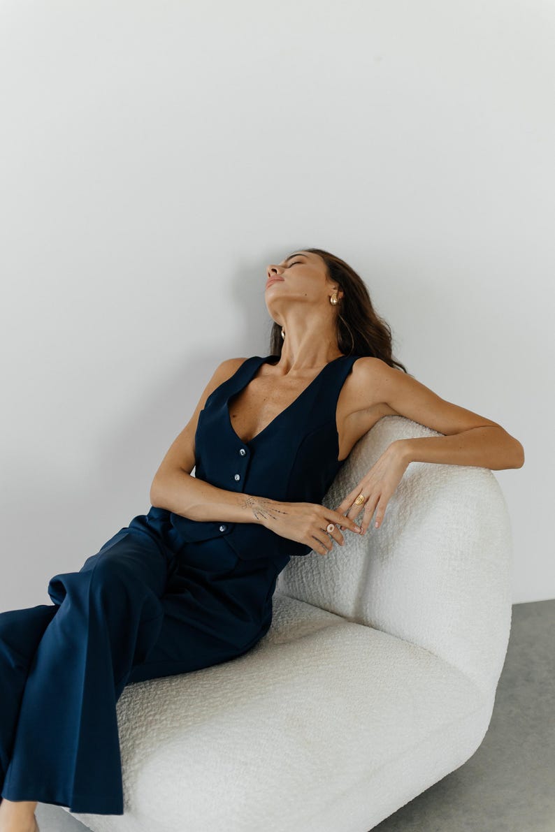 May include: A woman wearing a dark blue sleeveless vest and matching pants sits on a white fluffy chair. She has her arms crossed and is looking up.