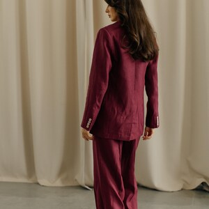 May include: A burgundy suit, consisting of a blazer and wide-leg pants. The blazer has buttoned cuffs. The outfit is made of a textured fabric. The person is standing in front of a neutral-colored curtain.
