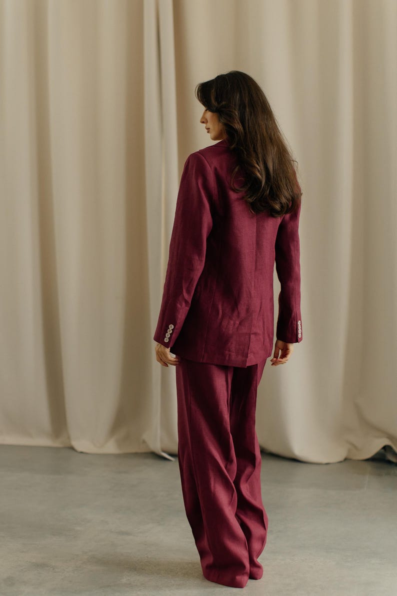 May include: A burgundy suit, consisting of a blazer and wide-leg pants. The blazer has buttoned cuffs. The outfit is made of a textured fabric. The person is standing in front of a neutral-colored curtain.