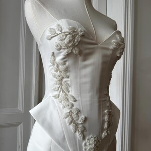 May include: A white satin corset top with a sweetheart neckline and a peplum waist. The bodice is adorned with beaded floral and leaf embellishments. The corset is displayed on a mannequin.
