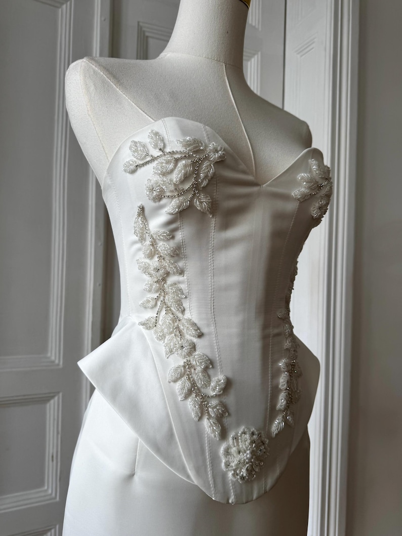 May include: A white satin corset top with a sweetheart neckline and a peplum waist. The bodice is adorned with beaded floral and leaf embellishments. The corset is displayed on a mannequin.