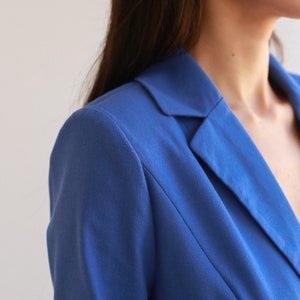 May include: Close-up of a blue blazer with a notched collar. The blazer is made of a smooth, textured fabric.
