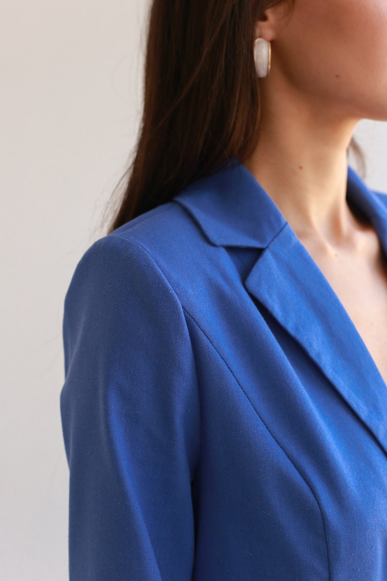May include: Close-up of a blue blazer with a notched collar. The blazer is made of a smooth, textured fabric.