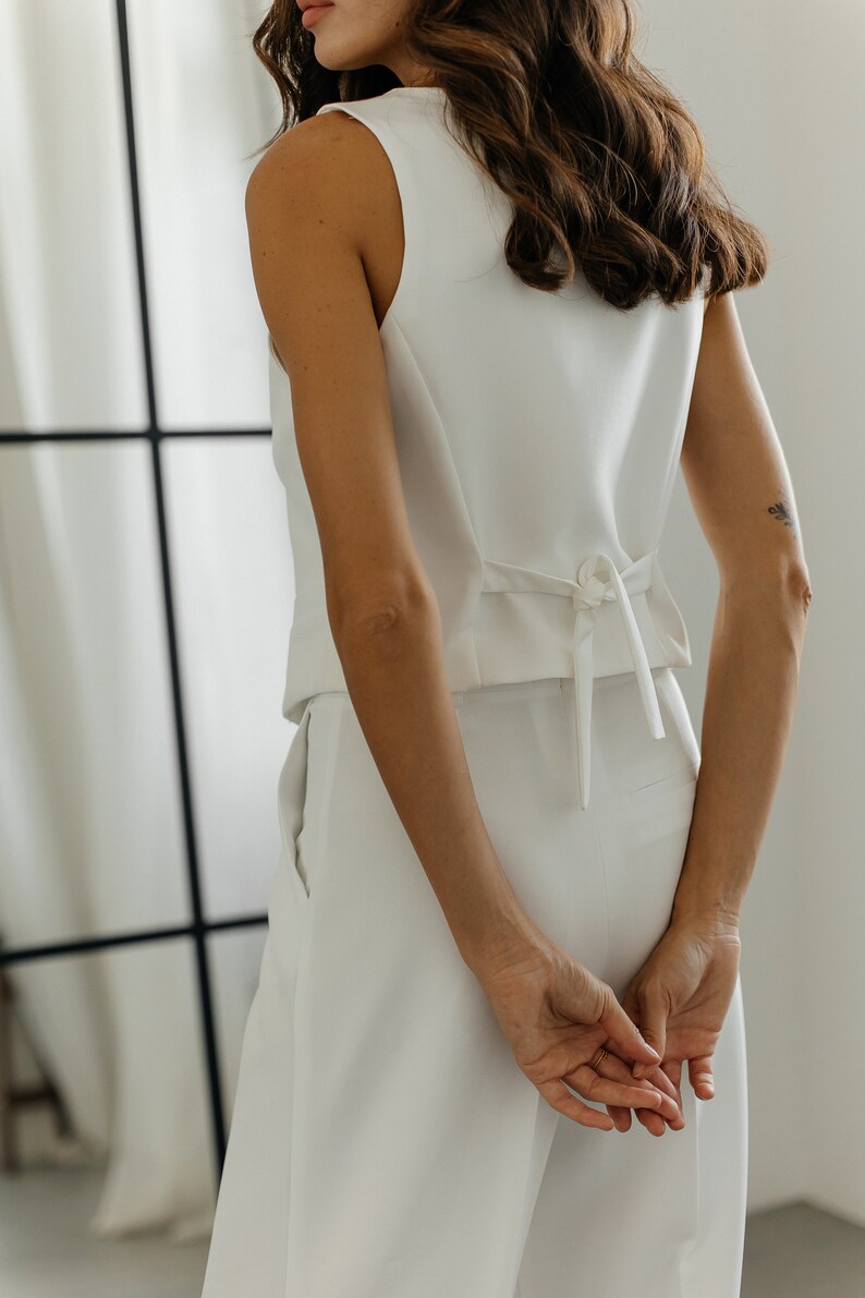 May include: A sleeveless white vest and matching pants. The vest has a tie detail at the back. The model's hands are clasped behind their back. The outfit is simple and elegant.