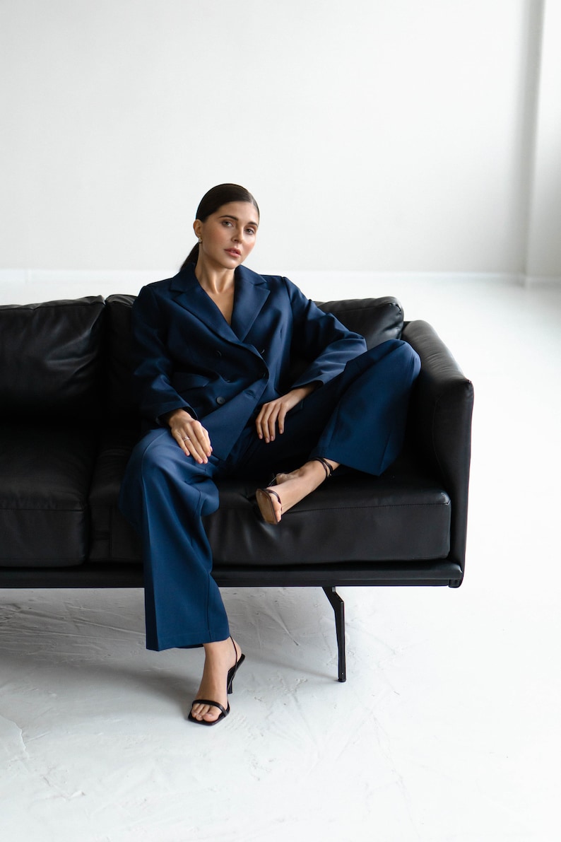May include: A woman wearing a dark blue suit sits on a black leather sofa. She is wearing black heels and has her hair pulled back.