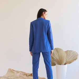 May include: A woman wearing a blue blazer and matching pants. She is standing in front of a white wall with a brown fabric on the floor.