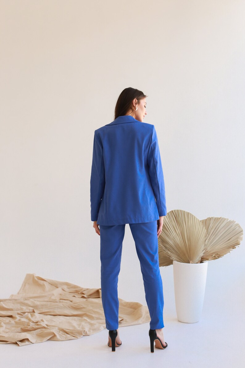 May include: A woman wearing a blue blazer and matching pants. She is standing in front of a white wall with a brown fabric on the floor.