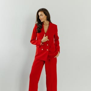 May include: A woman wearing a red double-breasted blazer and matching wide-leg pants. She is standing with her hands in her pockets and looking to the side.