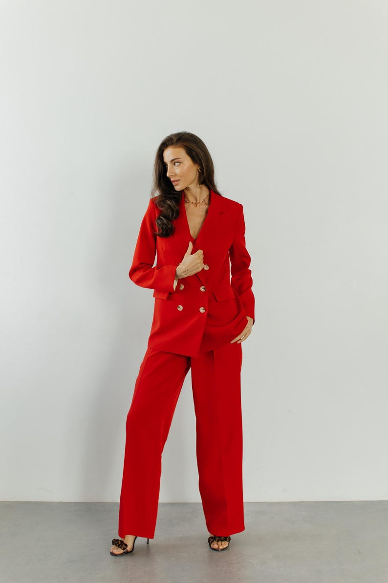 May include: A woman wearing a red double-breasted blazer and matching wide-leg pants. She is standing with her hands in her pockets and looking to the side.