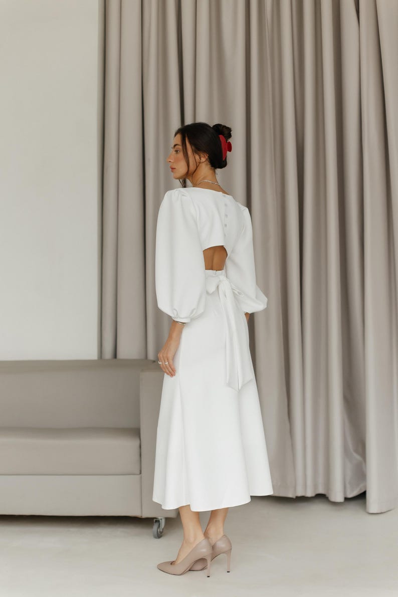 May include: A white midi dress with a low back and a bow tie detail. The dress has long sleeves with a puffed shoulder design. The model is wearing the dress with nude heels.
