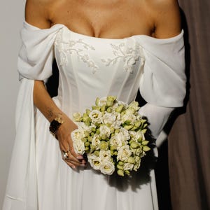 May include: A white, off-the-shoulder wedding dress with a fitted bodice and a flowing skirt. The dress has delicate floral embroidery and is paired with a bouquet of white and green roses. The sleeves are long and flowing.