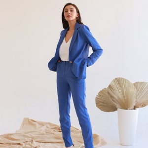 May include: A woman wearing a blue blazer and matching pants with a white v-neck shirt. She is standing on a beige fabric surface and wearing black heels.