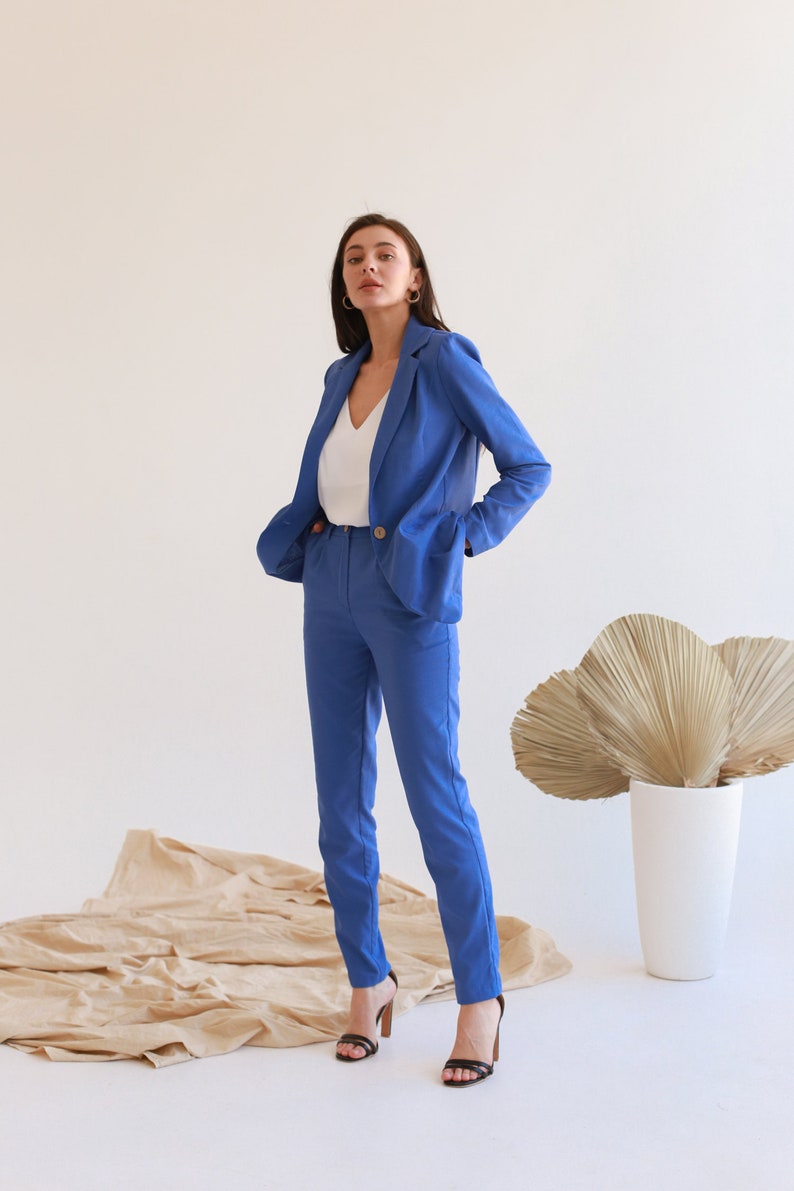 May include: A woman wearing a blue blazer and matching pants with a white v-neck shirt. She is standing on a beige fabric surface and wearing black heels.
