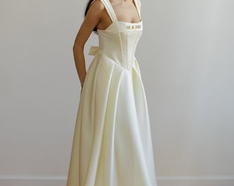 May include: A white satin wedding dress with a fitted bodice and a full skirt. The bodice features a square neckline and a corset-style design with a bow at the back.