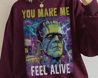 May include: Maroon sweatshirt with a graphic of Frankenstein's monster and the text "YOU MAKE ME FEEL ALIVE." The design features a green, blue, and purple color scheme. The sweatshirt is a solid maroon color.