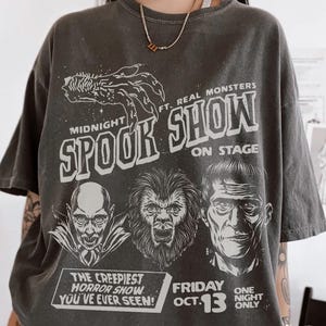 May include: Dark grey t-shirt with a graphic promoting the "Midnight Spook Show" featuring classic horror monsters. The design includes the text "The creepiest horror show you've ever seen!" and "Friday Oct. 13".