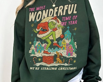 Può includere: Una felpa girocollo verde scuro con una stampa grafica di un personaggio verde in costume da Babbo Natale su un camino. Il testo recita THE MOST WONDERFUL TIME OF THE YEAR e WE'RE STEALING CHRISTMAS sopra e sotto l'illustrazione.