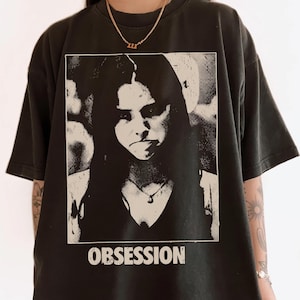May include: A black short-sleeve t-shirt featuring a rectangular graphic print of a person with long dark hair. The print is in a high-contrast black and white style. Below the image, the word OBSESSION is printed in bold white capital letters.