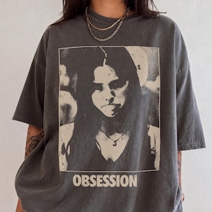 May include: A charcoal gray short-sleeve t-shirt featuring a large, rectangular, off-white graphic print of a person's face with long dark hair. Below the portrait, the word "OBSESSION" is printed in a bold, sans-serif, off-white font.