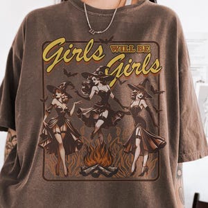 Puede incluir: Camiseta marrón con el texto "Girls Will Be Girls" en un diseño amarillo y marrón. El gráfico presenta tres brujas bailando alrededor de una hoguera. La camiseta es de gran tamaño y tiene mangas cortas.