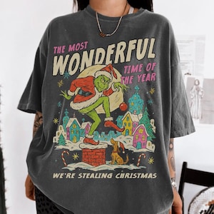 May include: Dark gray t-shirt with a Christmas-themed graphic. The design features a green character in a Santa hat, a dog, and colorful houses. Text on the shirt reads "THE MOST WONDERFUL TIME OF THE YEAR" and "WE'RE STEALING CHRISTMAS."