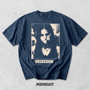 May include: A navy blue short-sleeve t-shirt featuring a rectangular graphic print of a person with long hair wearing a necklace. The word OBSESSION is printed in white capital letters below the image, and the word MIDNIGHT is below that.