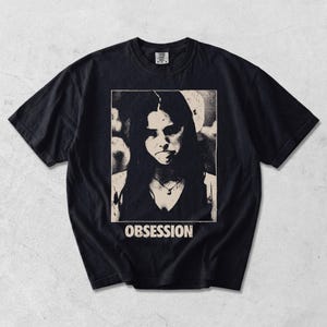 May include: A black short-sleeve t-shirt featuring a rectangular graphic print on the chest. The print displays a high-contrast, grainy black and cream portrait of a person with long hair and a necklace, with the word OBSESSION printed below.
