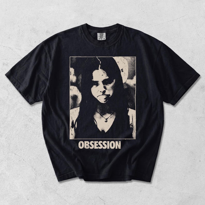May include: A black short-sleeve t-shirt featuring a rectangular graphic print on the chest. The print displays a high-contrast, grainy black and cream portrait of a person with long hair and a necklace, with the word OBSESSION printed below.