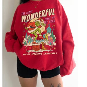 May include: Red sweatshirt with a graphic of the Grinch stealing Christmas. The design includes the text "The Most Wonderful Time of the Year" and "We're Stealing Christmas". The Grinch is depicted in a Santa hat.
