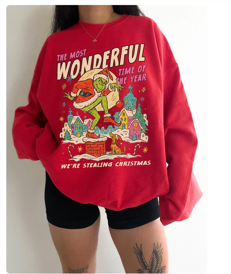 May include: Red sweatshirt with a graphic of the Grinch stealing Christmas. The design includes the text "The Most Wonderful Time of the Year" and "We're Stealing Christmas". The Grinch is depicted in a Santa hat.