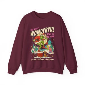 May include: Burgundy sweatshirt with a graphic featuring a green character in a Santa hat, standing on a chimney. The text reads "The Most Wonderful Time of the Year" and "We're Stealing Christmas."