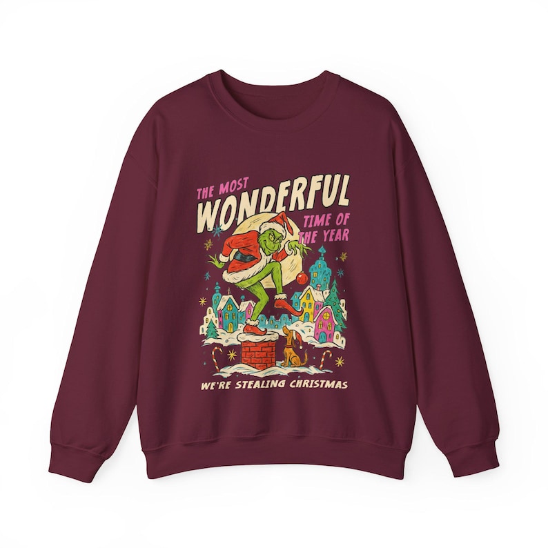 May include: Burgundy sweatshirt with a graphic featuring a green character in a Santa hat, standing on a chimney. The text reads "The Most Wonderful Time of the Year" and "We're Stealing Christmas."