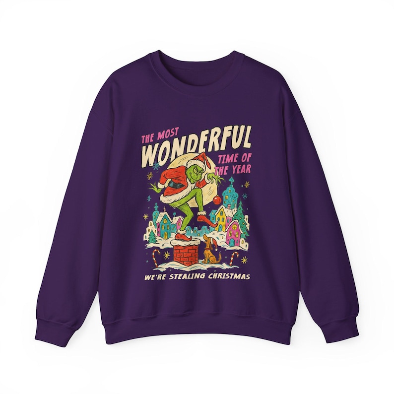 May include: A purple sweatshirt with a graphic of a green character in a Santa suit, standing on a chimney. The text reads "THE MOST WONDERFUL TIME OF THE YEAR" and "WE'RE STEALING CHRISTMAS". The design includes colorful houses and a dog.