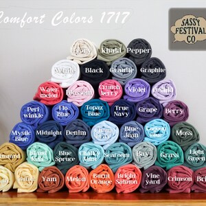 May include: A large pyramid-shaped stack of rolled t-shirts in various colors, labeled Comfort Colors 1717 and Sassy Festival Co. The shirts are arranged in rows, displaying a wide spectrum of shades including ivory, black, blue, green, and red tones.