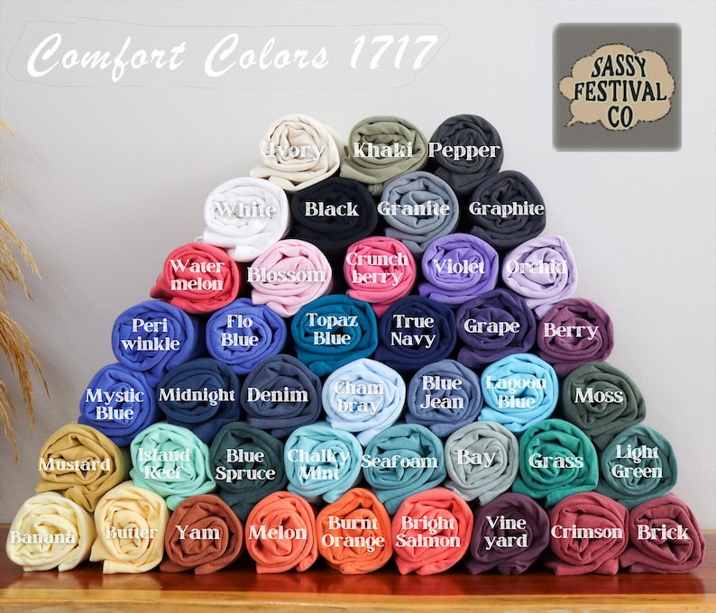 May include: A large pyramid-shaped stack of rolled t-shirts in various colors, labeled Comfort Colors 1717 and Sassy Festival Co. The shirts are arranged in rows, displaying a wide spectrum of shades including ivory, black, blue, green, and red tones.