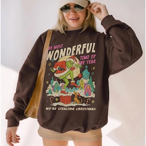 May include: Brown sweatshirt with a graphic of the Grinch stealing Christmas. The text on the sweatshirt reads "The Most Wonderful Time of the Year" and "We're Stealing Christmas". The Grinch is depicted in a Santa suit.