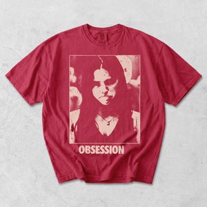 May include: A red short-sleeve t-shirt featuring a rectangular graphic print of a person with long hair wearing a necklace. The print is in a lighter shade of red and includes the word OBSESSION printed in capital letters below the image.
